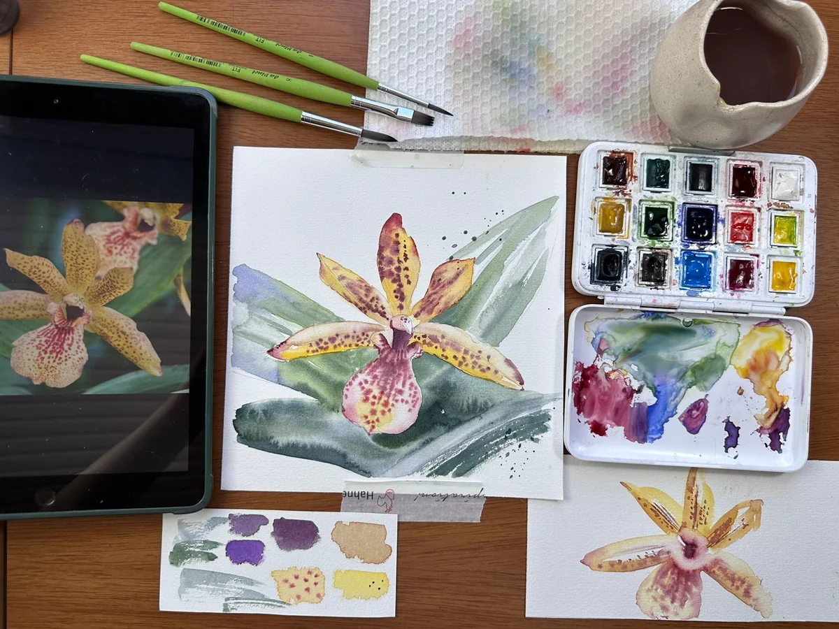 Mesa de trabalho da Carolina Spina com aquarela de orquídea amarela em processo, paleta de cores e pincéis